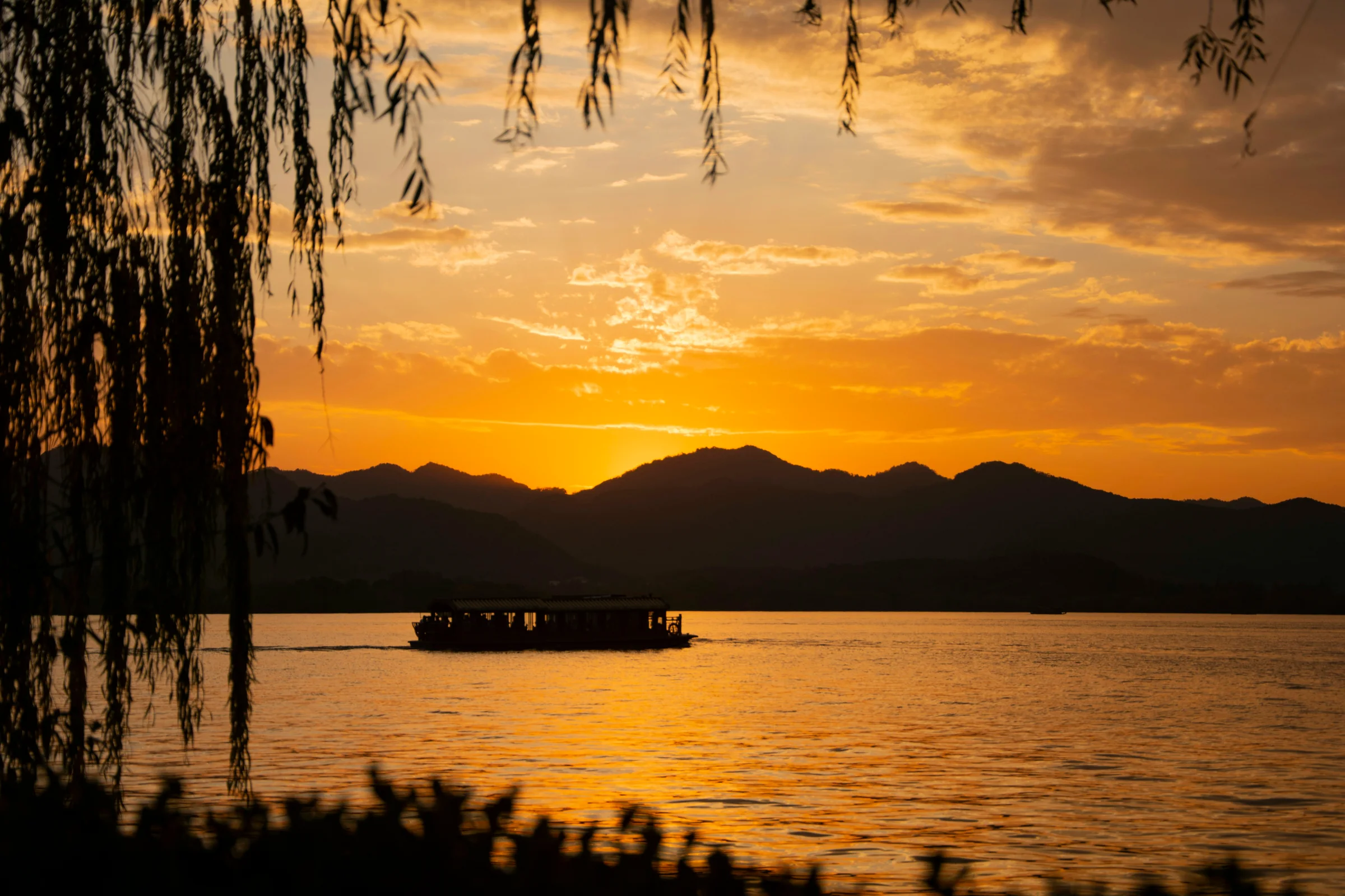 Willow branches frame a boat on Hangzhou’s West Lake, with golden clouds and layered hills beyond.