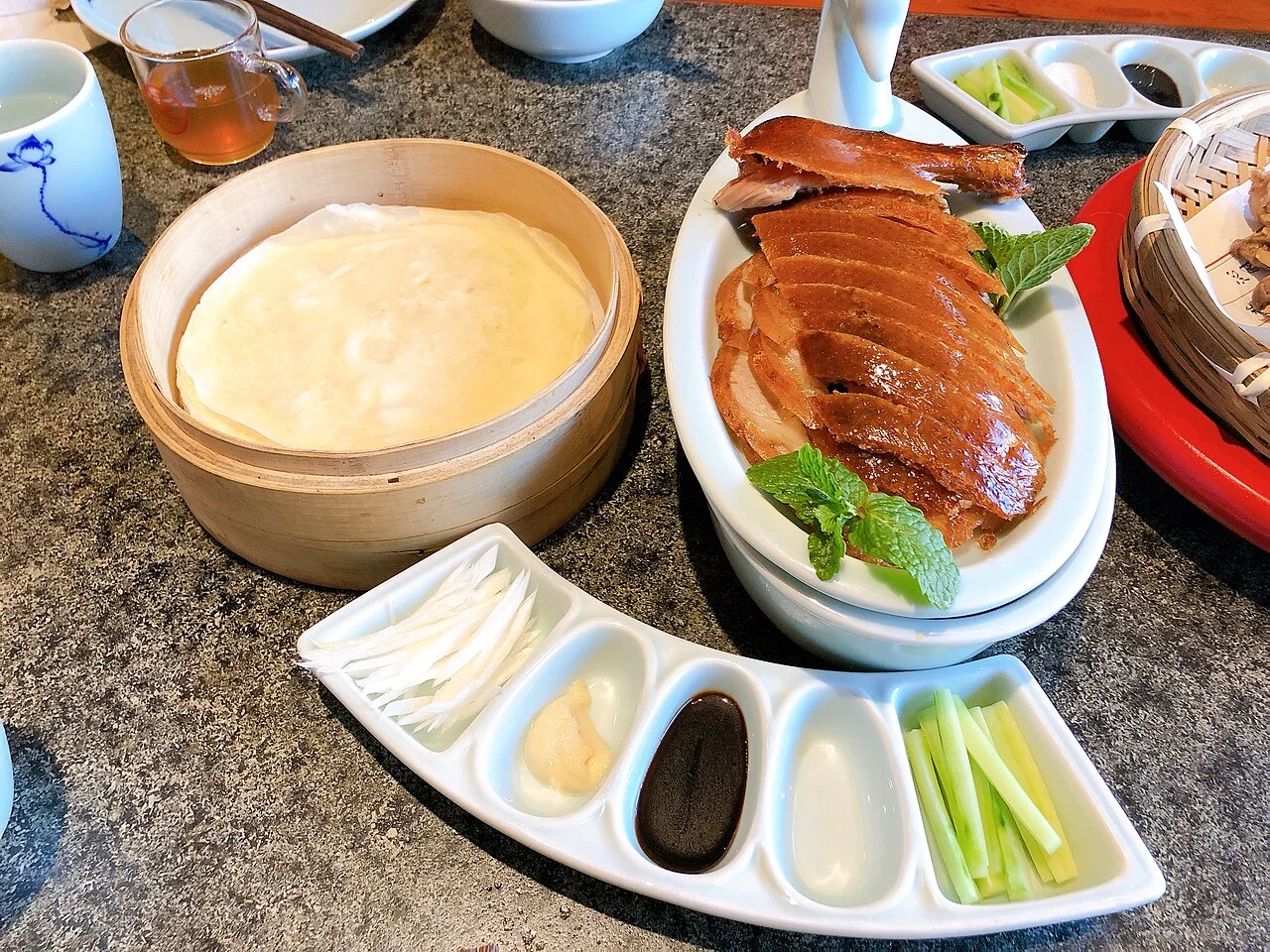 Sliced Peking duck on a white platter beside a bamboo steamer of pancakes, shredded scallion, cucumber and sauces.