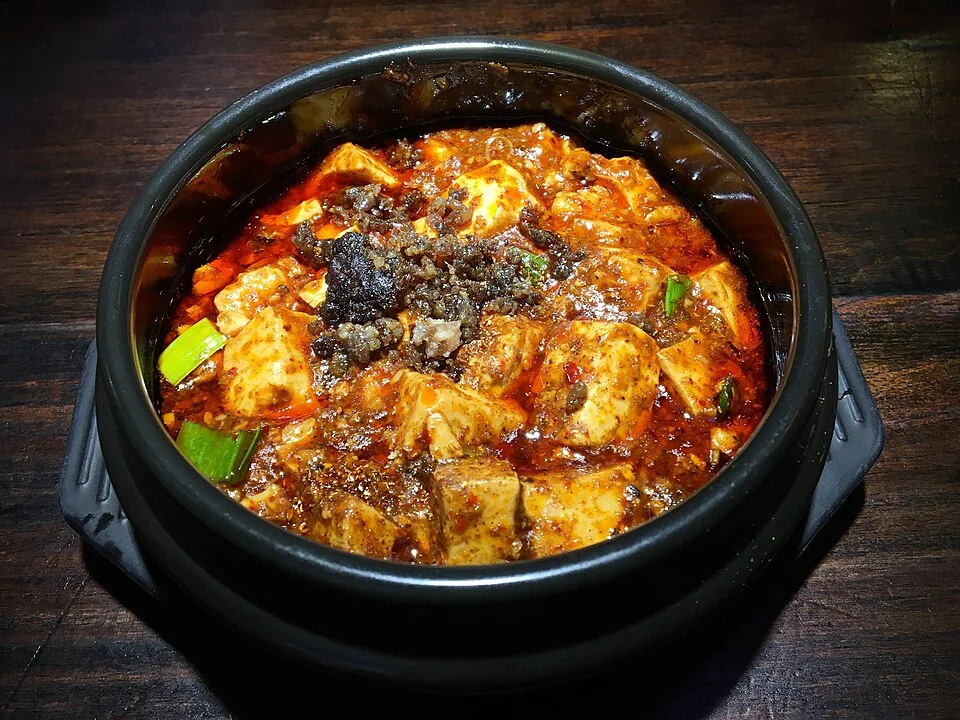 Mapo tofu in a black pot on a dark wooden table, with red chilli oil, minced meat and pieces of scallion.