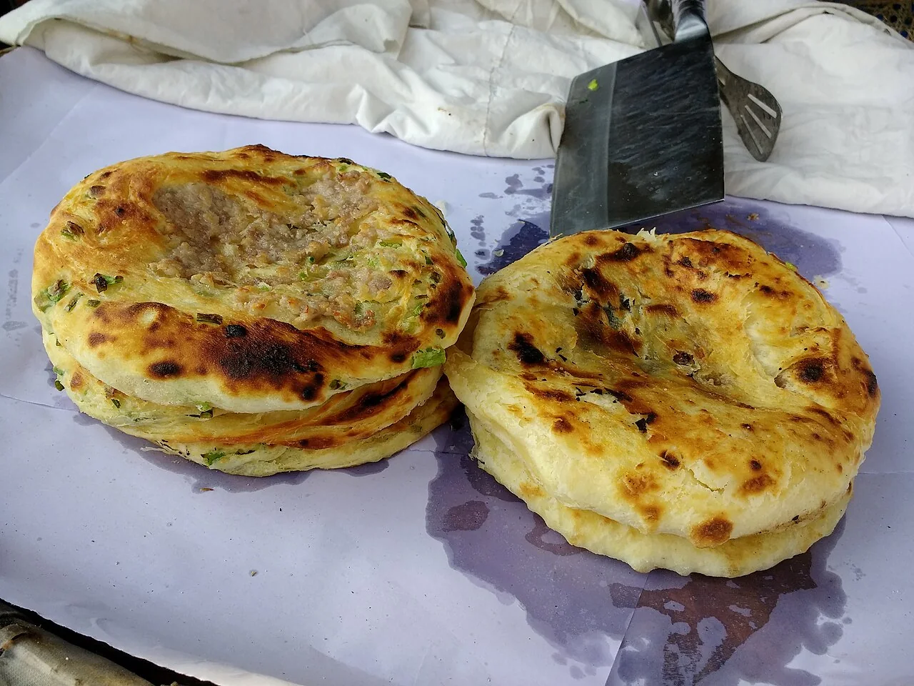 Golden meat-filled baba breads in Xizhou, with browned crusts, visible filling and chopped greens, on paper beside a metal spatula.