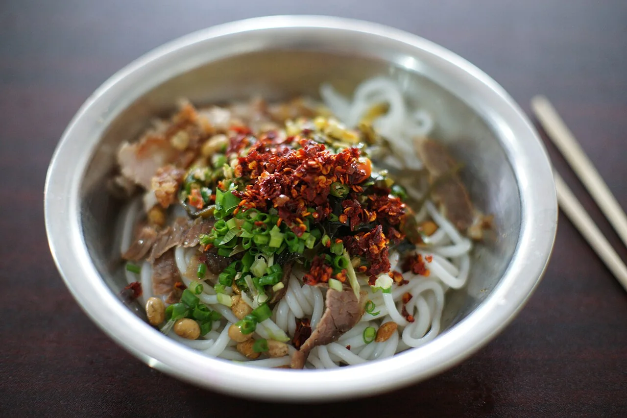 Guilin rice noodles in a metal bowl with sliced meat, chopped scallions, peanuts and red chilli, beside a pair of chopsticks.