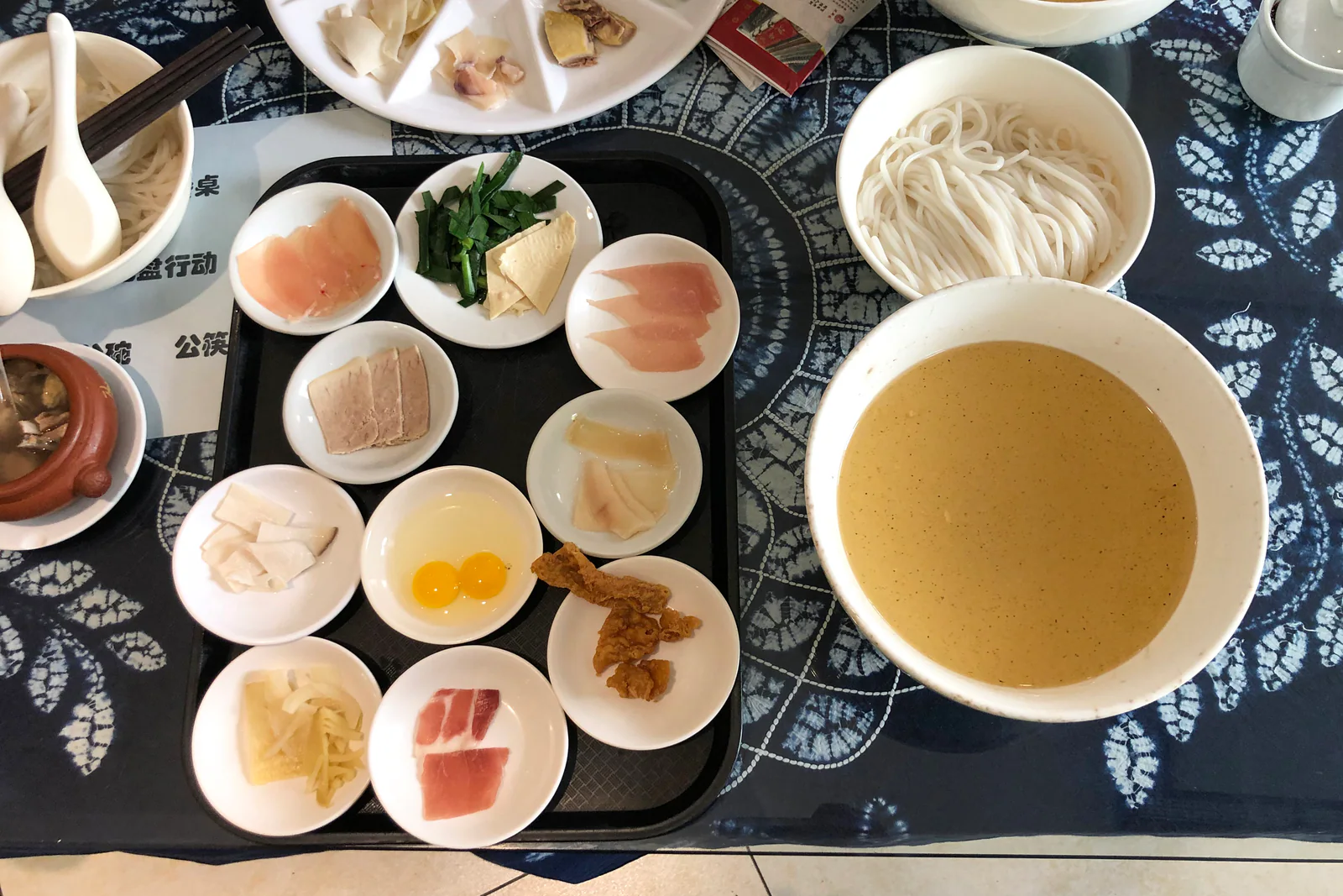Crossing-the-bridge noodles on blue-and-white patterned cloth, with a large bowl of broth, rice noodles and separately plated ingredients.