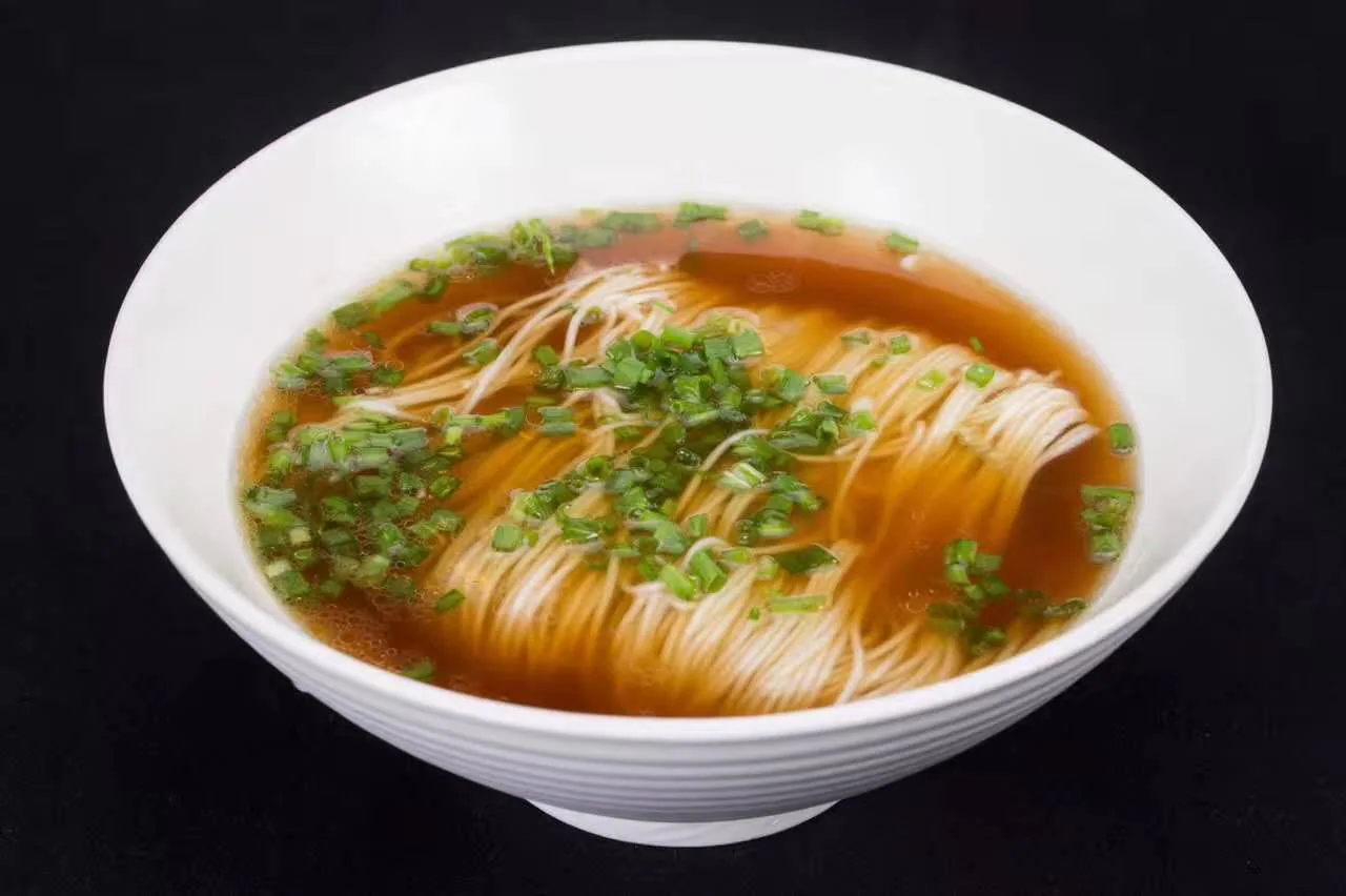 Fine Suzhou-style noodles in red broth, scattered with chopped scallions in a white bowl against a dark background.