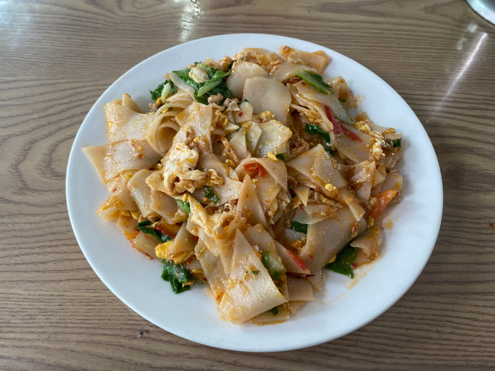 A white plate of stir-fried rice cake slices, egg and vegetables on a wooden table.
