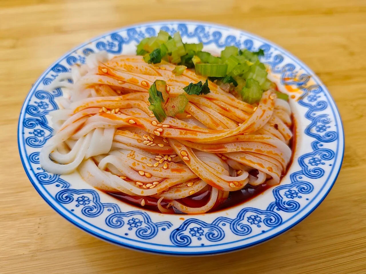 Broad ribbons of liangpi with red dressing and sesame seeds on a blue-and-white plate, topped with green vegetables in Xi’an.