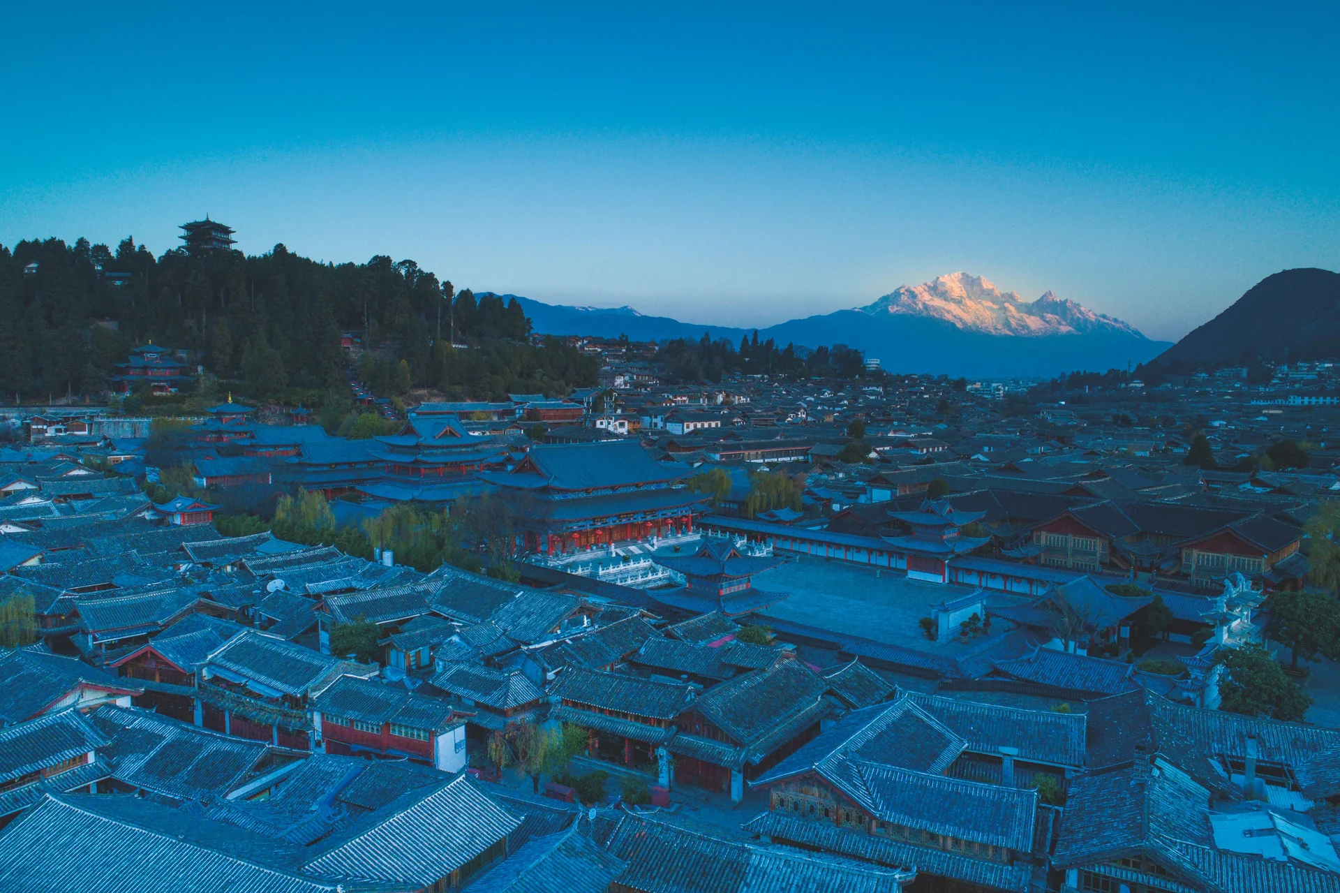 Layered tiled roofs of Lijiang Old Town at dusk, with warm light on a distant mountain peak.