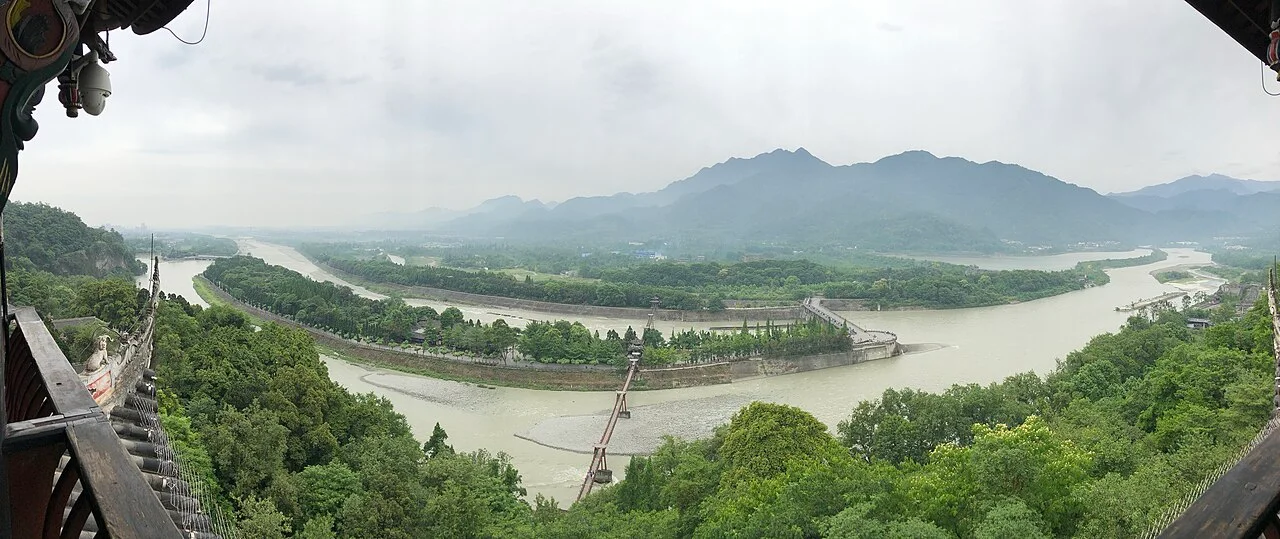 A panoramic view of Dujiangyan, with channels around wooded dividing embankments, a suspension bridge and mountains beyond.