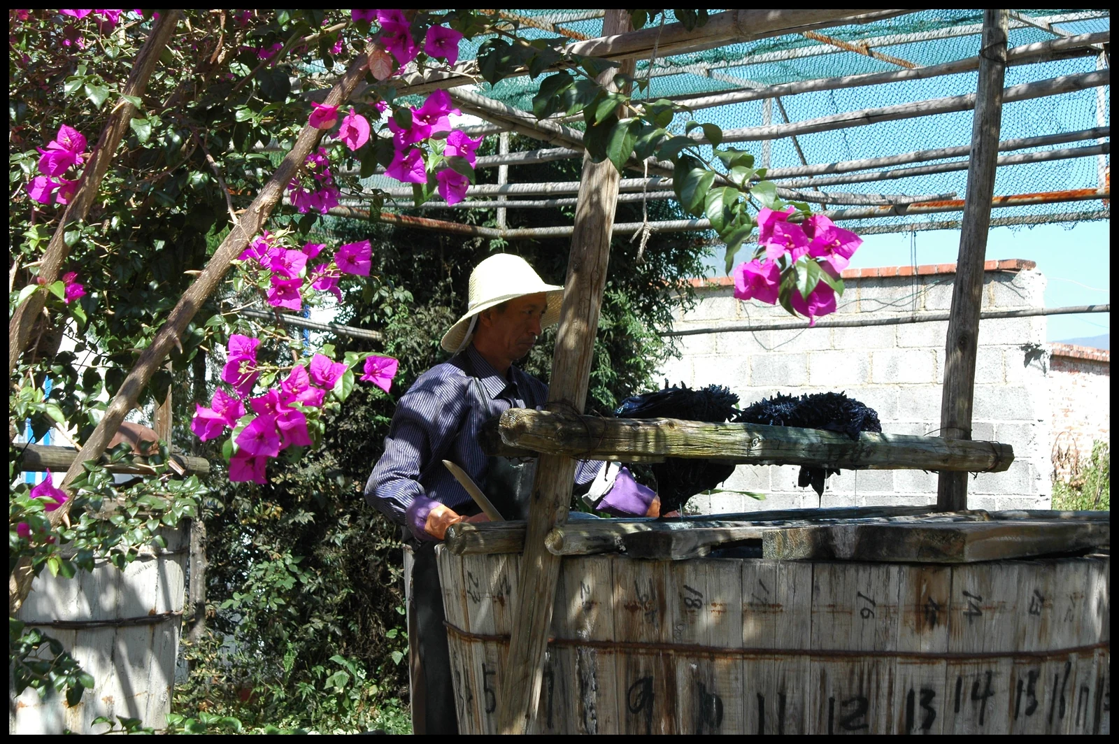 A worker beside a wooden dye vat in Dali, with bundles of dark blue tied cloth on a timber frame and flowers overhead.