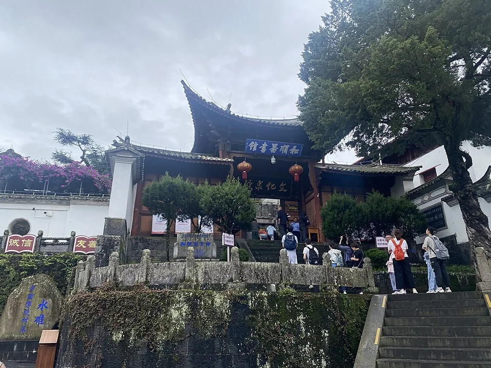 The entrance to Heshun Library in Tengchong, with a blue sign beneath tiled eaves, visitors on stone steps and a tall tree to the right.