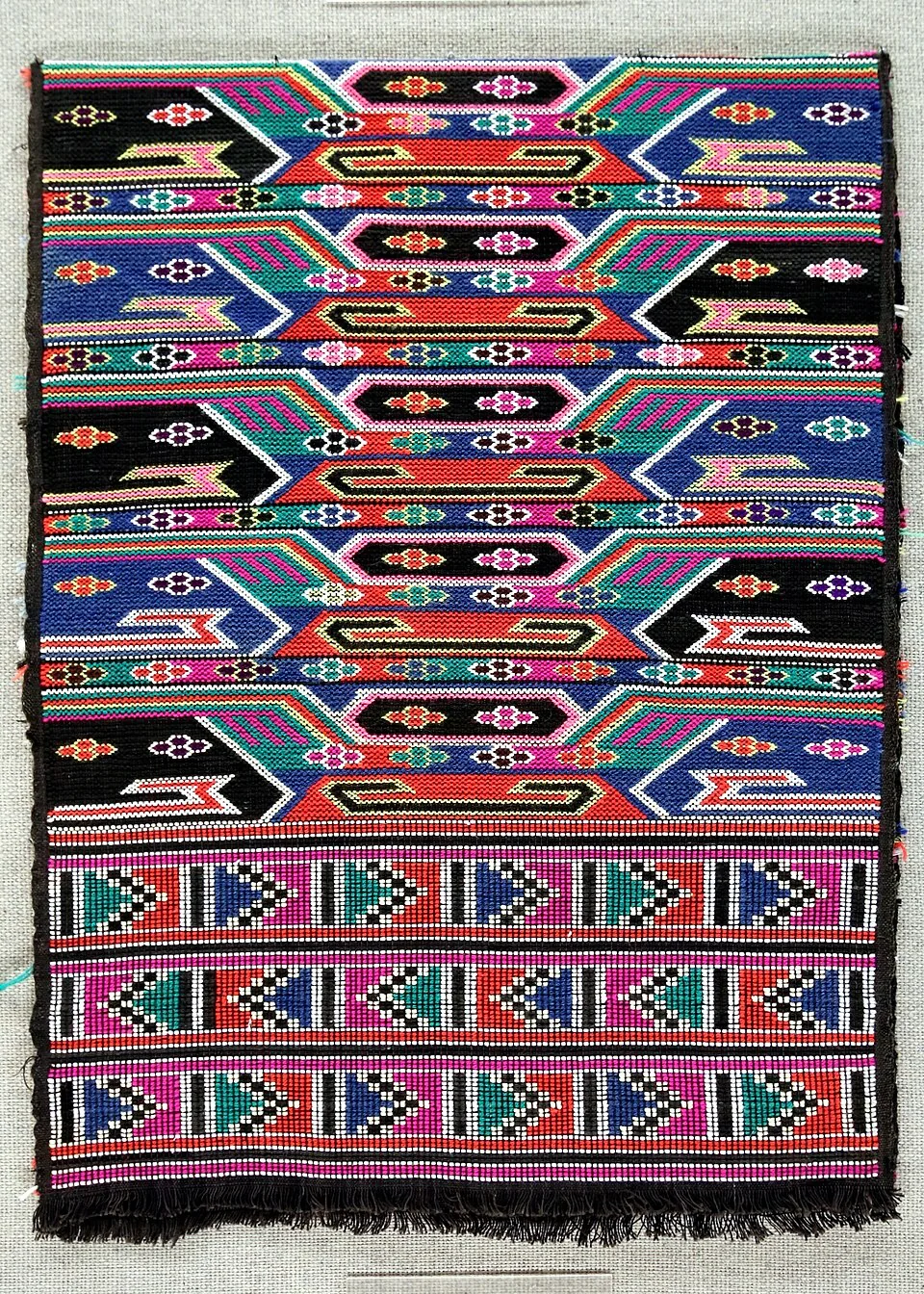 A Longshan Tujia brocade displayed at Shanghai Museum, with interwoven red, blue, green and black geometric patterns and a fringed edge.